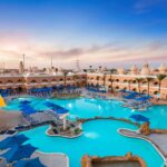 Weitläufige Poollandschaft mit blauen Liegen und Sonnenschirmen vor einem orientalisch gestalteten Hotel bei Sonnenuntergang.