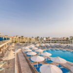 Großer Poolbereich mit weißen Sonnenschirmen, blauen Liegen und umliegender Hotelanlage im Pickalbatros Dana Beach Resort bei Sonnenuntergang.