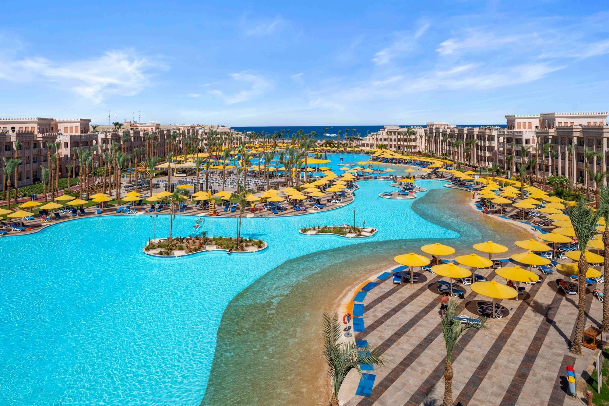 Große, geschwungene Poollandschaft mit gelben Sonnenschirmen, Palmen und Liegestühlen im Pickalbatros Palace Resort am Roten Meer.